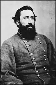 Black and white photo of a heavy-set, dark-haired and bearded man. He wears a gray military uniform with two rows of buttons.