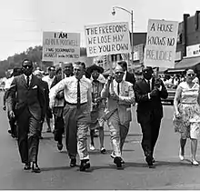 A group of four middle-aged men in suits and one woman in a dress walk in the first rank of a procession of individuals down the middle of a street. Brick upper stories of storefronts appear in the background, from middle to the right; tops of trees appear in the distance, far left. Three placards tacked onto pickets and held by two men in the second rank and one in the first rank read as follows. "I Am John A. Maxwell I Was Discriminated Against In The Pointes." "The Freedoms We Lose May Be Your Own." "A House Holds No Prejudice."