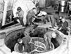 Soldiers and workmen, some wearing steel helmet, clamber over what looks like a giant manhole.