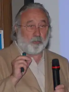 A portrait photograph of Bottazzo speaking while holding a microphone in his left hand and raising his right hand.