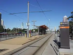 Island platform with a canopy in the centre. A shallow ramp leads up to the platform. At the top of the ramp an overhead wiring support mast is flanked by ticket validation machines on either side. A sign at the entrance to the stop directs passenger to buses or the beach.