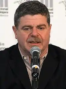 A 56-year-old man with dark gray hair talking into a microphone, looking at something beneath the camera.