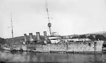 A four-funnelled warship lies at anchor in a harbour.