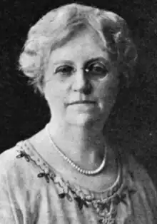 An older white woman, with coiffed white hair, wearing glasses, a strand of beads, and a light-colored dress with an embroidered round neckline