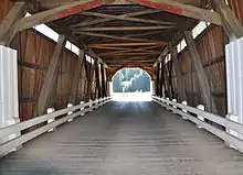 The view is lengthwise along the interior of a covered bridge. Its dark wooden supporting beams, wooden sides and deck, and wooden roof are exposed. Light shines through narrow rectangular openings near its roof. White metal guard rails run the length of the bridge on both sides. A wooded hill lies can be seen beyond the far end of the bridge.