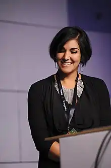 A woman looks downward from a podium before a purple backdrop.