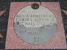 Moon Landing monument, with square pink terrazzo surround (not the usual charcoal color), with light gray terrazzo Moon disk showing TV emblem at top and the brass lettering "Neil A. Armstrong, Edwin E. Aldrin and Michael Collins, 7/20/69, Apollo XI"