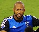 A man with a shaven head wearing a blue soccer jersey.