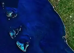 Deep blue ocean, with a large green land mass to the east, and four groups of islands to the west. Only the largest islands are easily distinguishable, but the island groups are rendered distinctive by the very light blue lagoons within them, and by the white lines of breakers marking their southern edges.