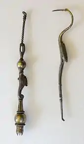 Two long, thin metal objects. The right object resembles a double-ended dentist pick in the shape of a crane bird. Its legs form the lower half, ending in a small pick; the body and wings form the handle in the middle; whereas the curved neck, head and long pointed beak form the upper pick. The left object is straight; a thin twisted rod ends in a large ball, followed by a fish motif again bounded by a ball, and ending in a crown with its peaks pointing inwards, presumably to hold bristles.