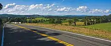 A rural landscape with a mix of fields and woodlots and low wooded mountains extends in the distance beneath a blue sky full of clouds. From bottom right to center left a paved road with a yellow stripe in the middle fills the image.