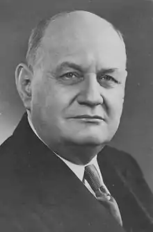 Black and white photograph of a man with a square face, strong chin and an under-chin, balding. He is looking into the middle distance, not directly at the camera, and is wearing a dark suit with a plain tie.