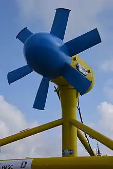 Looking up at the Sabella D10 turbine from ground level. At the top of the image are 6 large blades around a bulbous hub, all painted bright blue. The cylindrical nacelle behind is supported on a pile with tubular steel bracing forming a tripod. The Nacelle and foundation are painted bright blue.