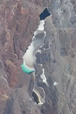 Space image of a white plain embedded in mountainous terrain, with a brownish lake to the south. A green lake lies in the middle directly next to the white area, while to the north lies a black lake