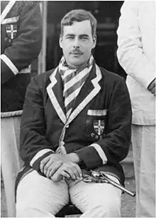 Black-and-white photograph of a young moustachioed man in a purple blazer, with a revolver on his lap.