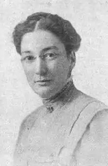 White woman wearing glasses and a high-necked blouse; her dark hair is parted at center and braided across the crown.