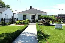 The single-story house has white walls, two windows, a central white door with a black door frame, and a black roof.