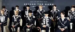 Eleven members of JO1 sitting in two rows in front of a backdrop of JO1 1st Asia Tour Beyond the Dark Limited Edition