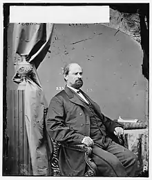 A man with dark hair, a mustache, and a long beard wearing a black jacket and vest and white shirt, sitting in a chair and facing right