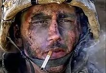 A man with dirt and ash on his face in a military helmet, staring intently forward while he smokes a cigarette