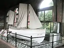 White-hulled small boat sitting on a base of stones behind a rail, within a museum. The name "James Caird" is visible. A stuffed penguin in a glass case stands nearby.