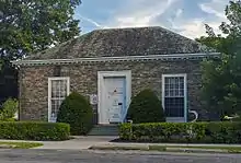 A light brown one-story three-bay stone building with a hipped shingled roof and mature oak trees on either side. On its front door a white sign reads "Hyde Park Free Library"