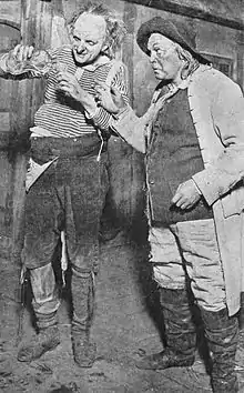 Two men wearing ragged clothing stand next to each other. Both are looking at the drink that one is pouring.