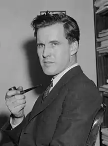 A young man wearing a suit and tie turns profile to the camera, holding a pipe in his hand.