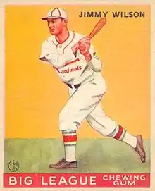 A baseball card image of a man wearing a white baseball uniform and cap with red trim swinging a baseball bat