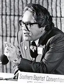 A man, Jimmy Allen, wearing a suit and tie sits at a table. He is mid-sentence and is pointedly gesturing at something out of frame.