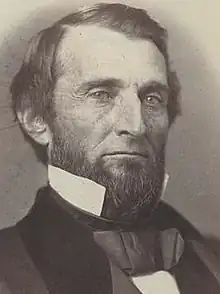 A man with black hair and a black beard wearing a black jacket and tie and white shirt