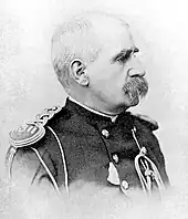 Profile of a balding white man with bushy, drooping mustache wearing an ornate military jacket with shoulder boards, shoulder cords, and a lanyard hanging from the chest.