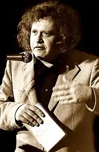 Van Doorn has curly hair and is speaking in a microphone, holding a notepad in his right hand, and making gestures with his left hand