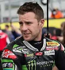 A man sporting a stubble is looking to the right of the camera and is wearing black and green motorcycling overall