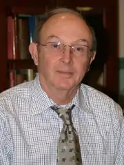 Elderly man balding with gray hair, wearing a gray suit jacket, white dress shirt, black necktie and tinted eyeglasses