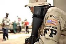 An Air Force officer, turned sideways in such a manner that his US flag patch is visible, carefully observes a group of Iraqi civilians carrying bags of sand whilst holding an armed weapon, on the lookout for any suspicious activity.