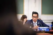 Khan is pictured listening to a witness testify. He wears a navy suit, gold and baby blue striped tie, and black glasses.