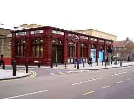 Single storey red glazed terracotta station building with tall arched windows. Glass shaded lamps project from the façade on ornate brackets and the words "Kilburn Park", "Entrance", "Exit" and "Underground" are displayed in black on a white moulded tile band above the window arches.