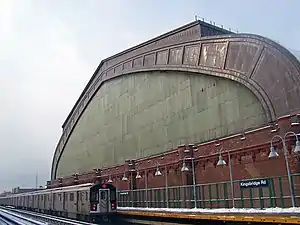 A train with an illuminated "4" in a circle on the front at a concrete platform with curved gray lights where a white-on-black sign reads "Kingsbridge Rd". Above it is a towering semicircular building with a greenish glass face trimmed in brown steel above brick with a castle-like roofline