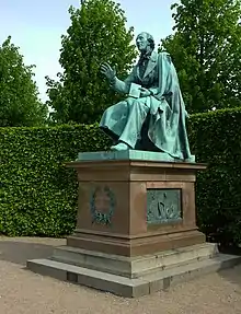 Bronze statue of a seated man raising his right hand while holding a book in his left, on a marble pedestal