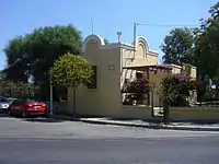 Durrell's house in Rhodes features Mediterranean architecture and has yellow-painted stucco or plaster walls. It is located on a paved asphalt street, with two cars parked parallel to it. The house is surrounded by several trees, shrubbery, roses, and flowering bushes.