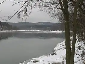 A grey lake under grey skies with snow and ice blocks on its shore and snow covered low mountains in the background