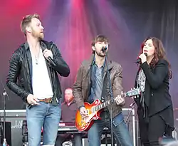 Two men and a woman performing on a stage, both singing into microphones.  All are holding microphones and one of the men is also playing a guitar.