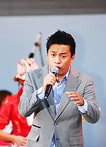 A picture of a brunette man performing on stage, he is wearing a coat with black-and-white polka dots, and a blue-striped shirt inside