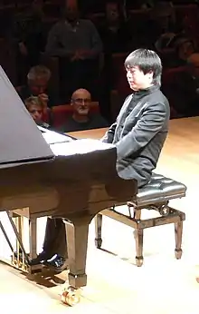 A Chinese man sits at piano, performing, while an audience watches.