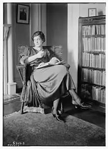 A white woman with dark hair, seated indoors, in front of a bookcase, with a book open in her lap