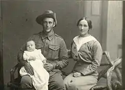 Black and white photograph of a seated man in military uniform holding a baby dressed in a white, flowing gown, with a woman sitting next to him. The woman has her arm around the man's back.