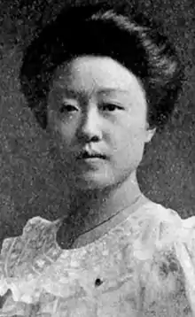 A young Chinese woman with her dark hair in a bouffant updo. She is wearing a light-colored dress with a lacy ruffled yoke.