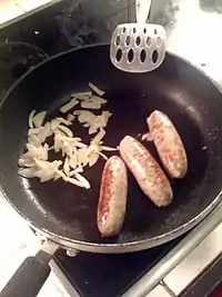 Three Lincolnshire sausages, with chopped onions in a frying pan with a spatula on a hob