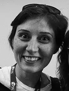 A candid grayscale portrait photo of a white woman; she is facing and smiling into the camera, and has a pair of spectacles resting on her short, dark hair.